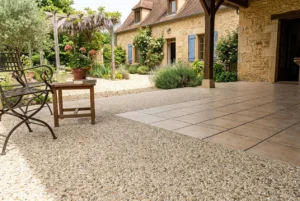 Allée et patio extérieurs avec pavage existant, entourés de végétation et d’une façade de maison en pierre; surface mixte : moquette de pierre sur carrelage près du coin repas.